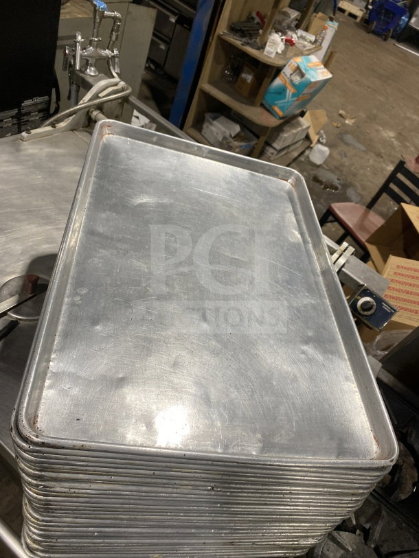 Stack of commercial stainless steel full-size baking sheet pans in used condition on industrial table.