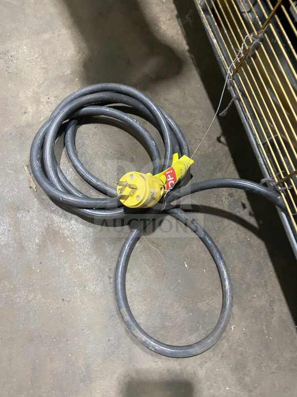Coiled electrical cable with a yellow industrial connector, resting on a concrete floor near a metal rack.