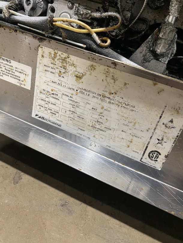 Southbend commercial stainless steel 6 burner range with 24" flat griddle, 2 full-size ovens, and poly wired rack on casters. Labels indicate installation and safety details.