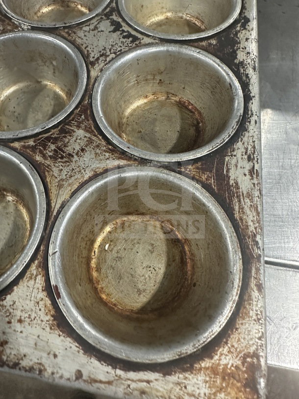 Commercial heavy-duty 24-cup stainless steel muffin pan, visibly worn, rust spots, suitable for baking large batches.