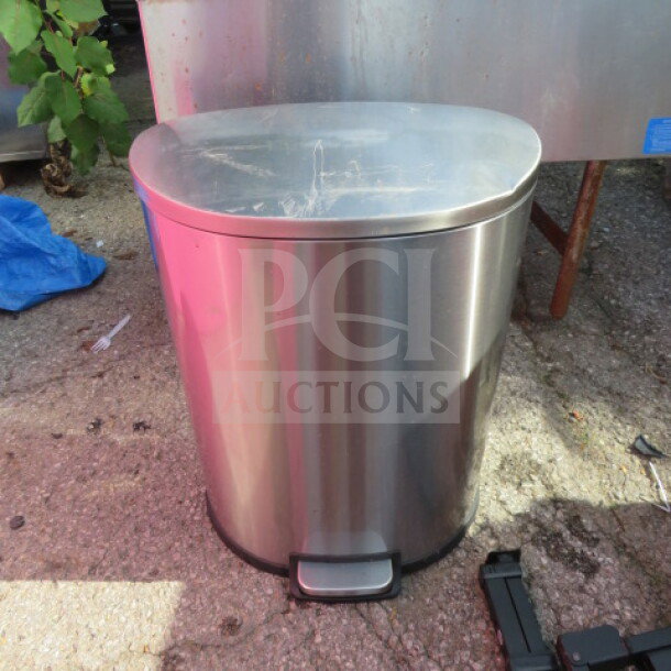 Stainless steel step trash can, oval shape, with manual pedal, visible surface wear, outdoor setting.