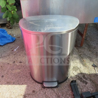 Stainless steel step trash can, oval shape, with manual pedal, visible surface wear, outdoor setting.