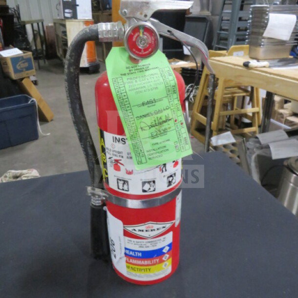 Amerex ABC fire extinguisher with tags showing inspection details. Red cylinder, silver nozzle, in used condition.