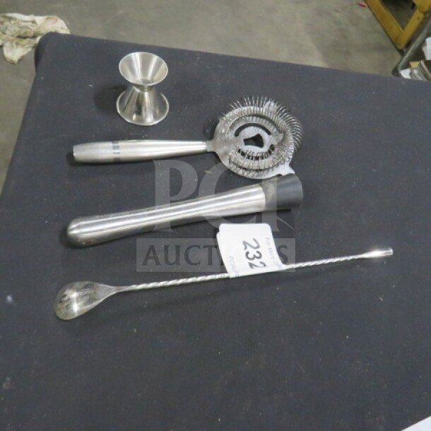 Assorted barware set including jigger, strainer, muddler, and stirrer on a black surface. Auction tag reads "232."
