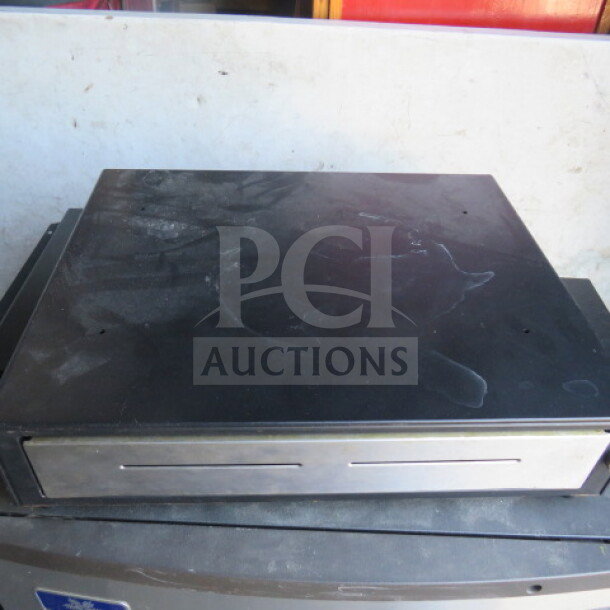 Black cash drawer with metal front, showing signs of wear. No visible brand or model markings. Auction item.