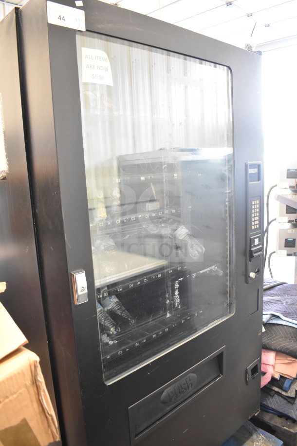 Seaga Commercial Floor Style Vending Machine, black with key lock, coin slot, and clear viewing panel. Sign reads "All items are now $0.50."