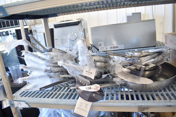 18 new Vigor SS1 Series stainless steel fry pans, various sizes, tags visible, on metal shelf, scratch and dent condition.