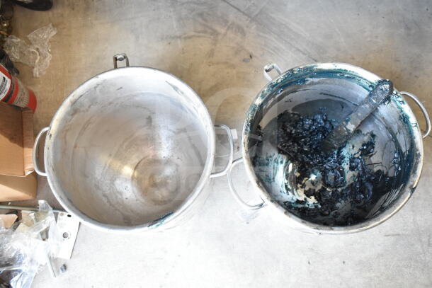 Two large metal mixing bowls, one clean, one with dark residue inside. Both have handles, industrial use.