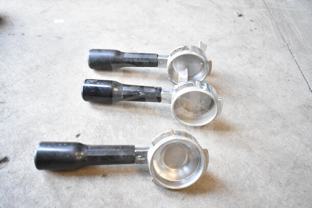 Three metal portafilters with black handles on a concrete surface. Part of a 7-item auction lot.