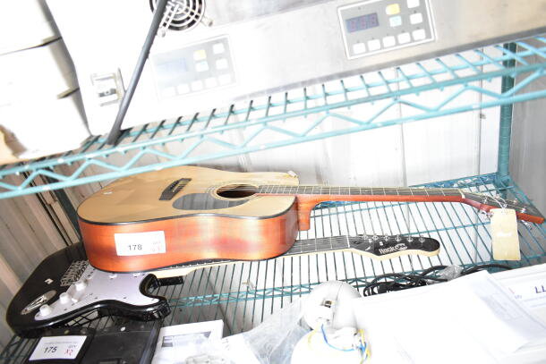 Two guitars on a metal shelf: a Cort acoustic with a tag and a RockJam electric, both in good condition.