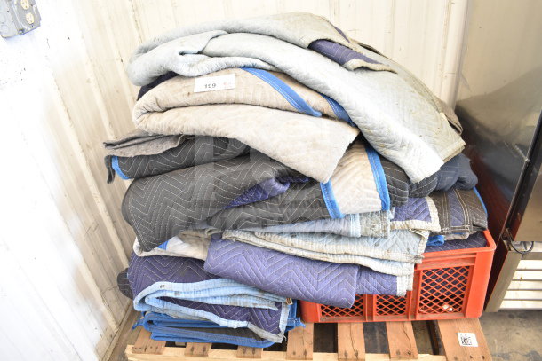 Pile of 14 moving blankets in mixed colors and patterns on a crate. Well-used condition, ideal for protecting items during transport.
