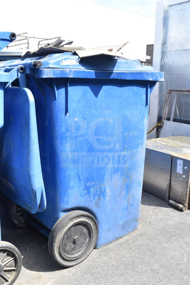 Two large blue poly trash cans with wheels, used condition, suitable for outdoor waste storage.