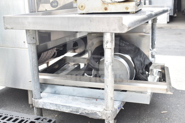 Stainless steel table with under shelf holding baking pans, signs of wear on legs, sturdy industrial design.