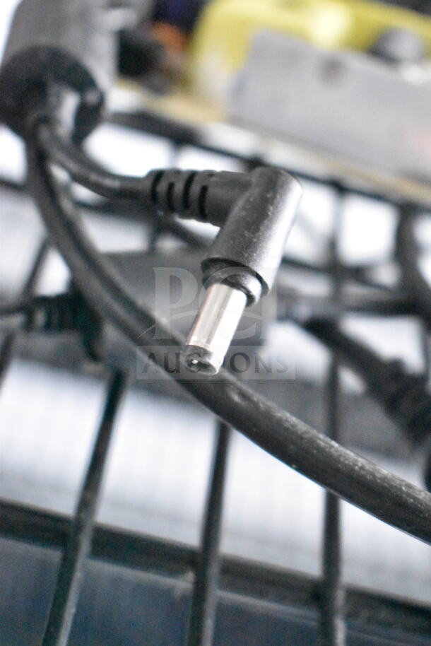 The image shows a close-up of a black electronic cable with connectors, placed against a blurred industrial background.