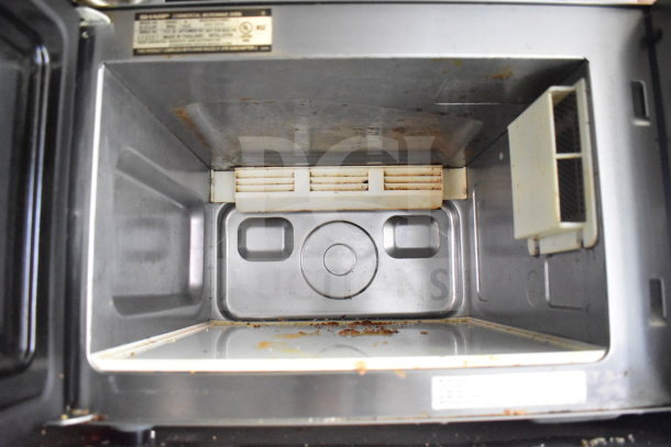Interior view of a used commercial microwave oven, showing slight wear and food residue. Manufacturer label visible.