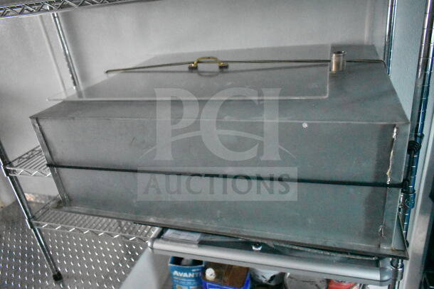 Metal storage box with a latch and pipe fitting, placed on a wire shelf. Condition appears used, with visible wear.