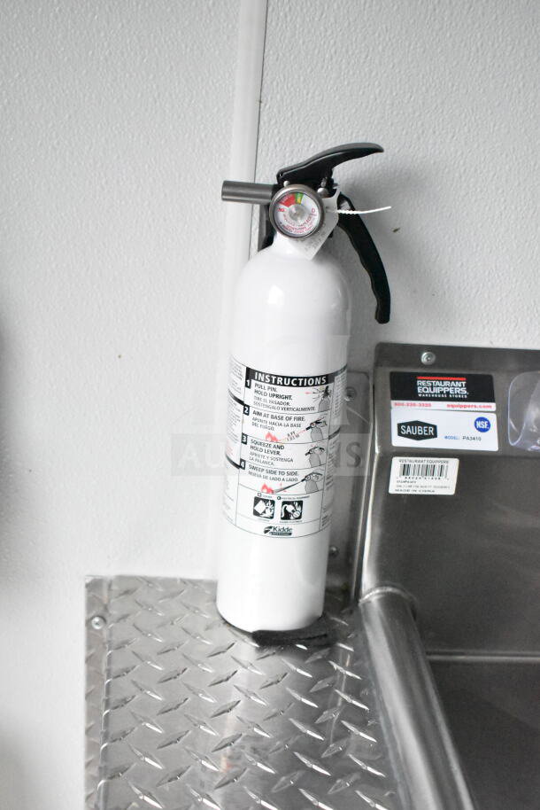 White Kidde fire extinguisher with instruction label mounted on diamond plate surface. Nearby Sauber model PA3410 tag.