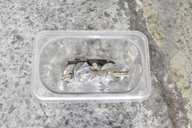 Plastic container with assorted metal components, screws, and a bracket on a textured concrete surface. Components appear used.