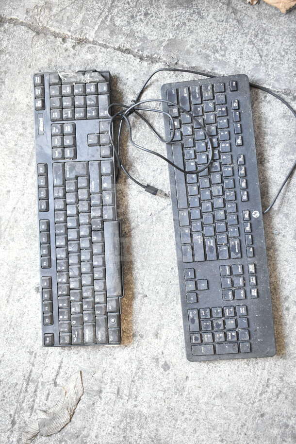 Two used black keyboards with USB cables on a concrete surface, showing signs of wear and dust.
