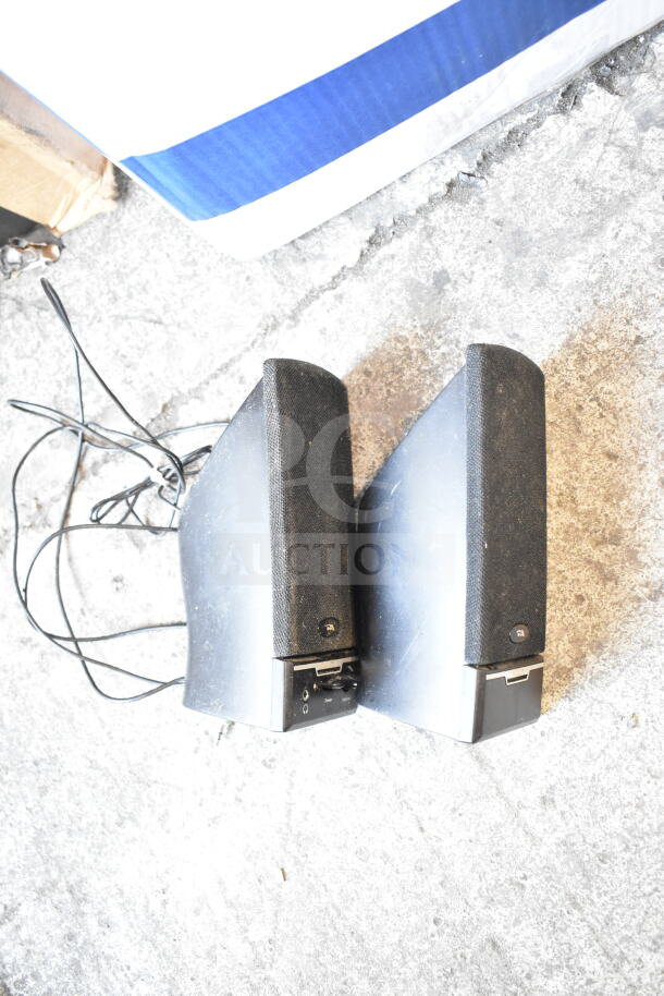 Pair of black wired speakers on a concrete surface, showing power and input ports, slightly worn condition.