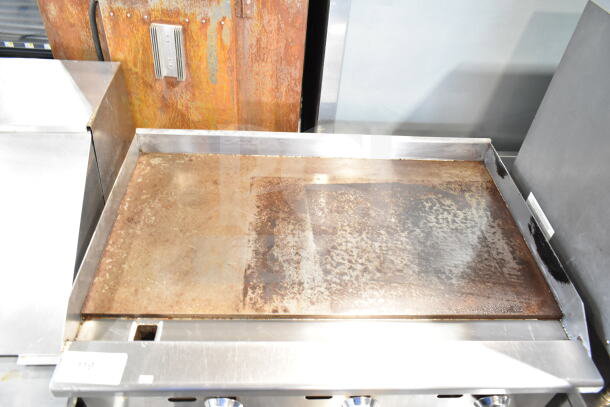 Commercial gas-powered countertop griddle, stainless steel, flat top, used condition, metal control knobs visible.