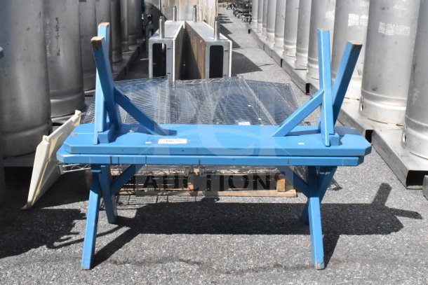 Blue wooden bench, upside down, with visible wear on legs, placed outdoors among metal containers. Tags on surface suggest auction details.