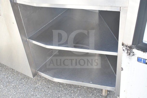 Stainless steel counter with two shelves, featuring a bay and sneeze guard. Visible wear on surfaces, MB label present.