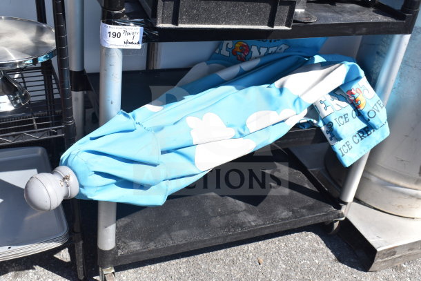 Black poly 2-tier cart with commercial casters, contains a folded blue umbrella with white cloud design. Tag reads "190/Qty 1."