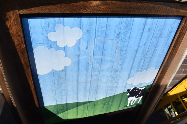 Tabletop with cow painting features a whimsical design of a cow, clouds, and field on a blue sky background. Wood frame, vibrant colors.