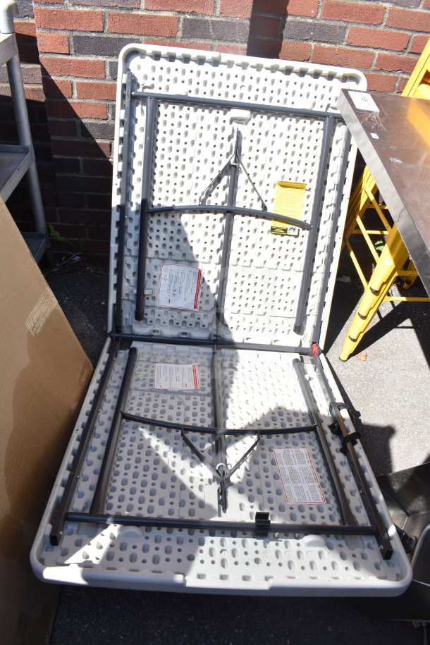 Bi Fold Folding Table, white plastic with metal legs, open and collapsed for storage. Visible warning labels, outdoor condition.