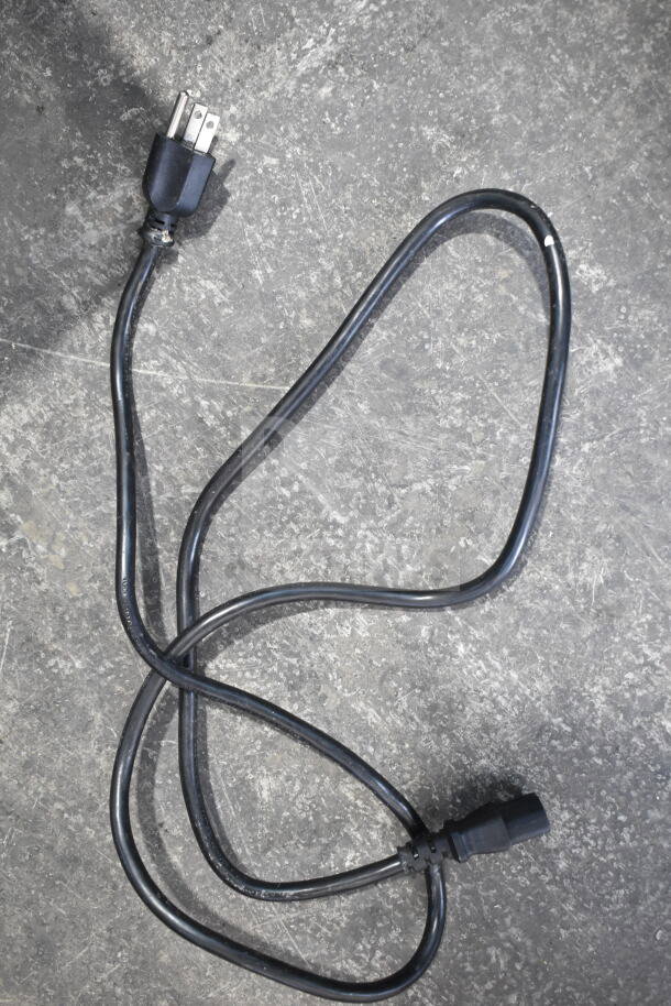 Black electrical cord on a concrete surface, featuring a standard plug and connector ends, in used condition.
