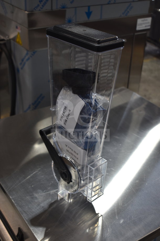 Brand new dry goods dispenser with clear container and black lid, marked with part number and revision label, scratch and dent condition.