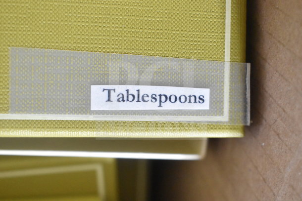 Box labeled "Tablespoons" on textured yellow surface, part of a set of new scratch and dent cutlery, including spoons and forks.