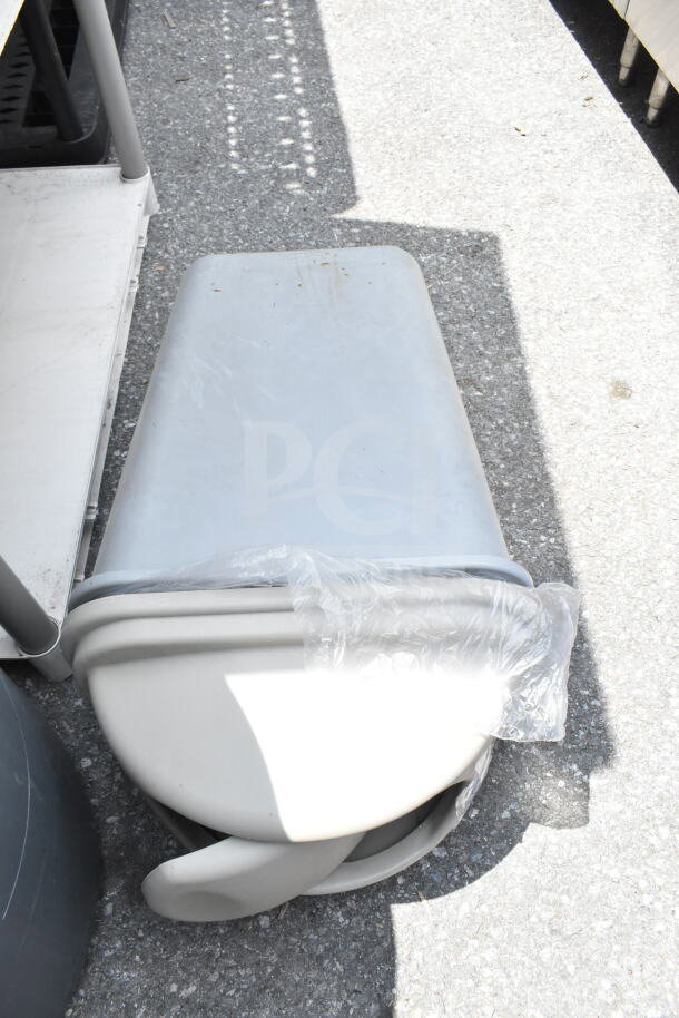 Two gray plastic trash cans stacked, outdoor setting, one wrapped in plastic, used condition.