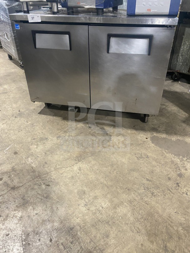 Two-door stainless steel undercounter refrigerator or freezer, commercial, maintained condition, with handles and wheels.