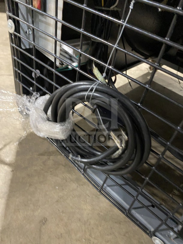 Coiled electrical cord attached to the back of a Dukers GDM40BS refrigerator, seen through metal grid casing.