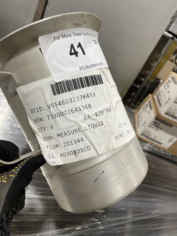 Brand new stainless steel liquid measuring cup with label showing item details including quantity, NSN, and price.