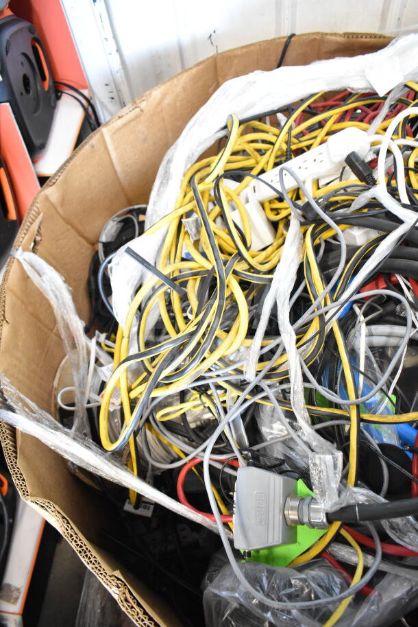 Lot of various cables in a cardboard box, including power strips and extension cords, mixed condition.
