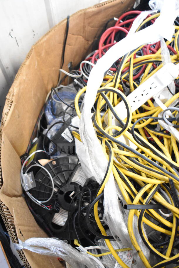 Lot of assorted cables in cardboard box, featuring various colors and types, some with labels and plastic wrapping.