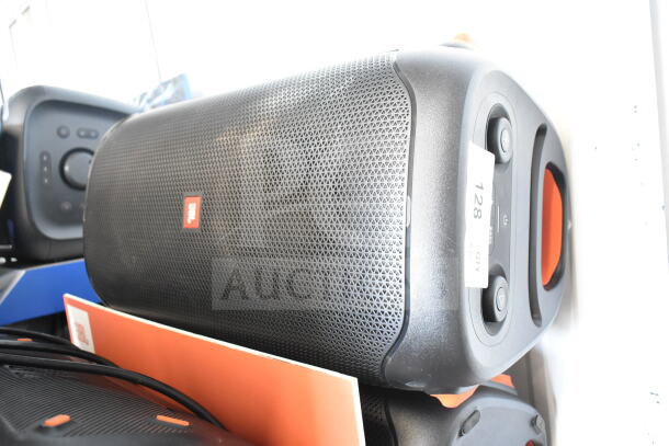 JBL speaker on store display, black grille, orange accents, slight wear. Visible tag reads "128 QTY," indicating stock number.