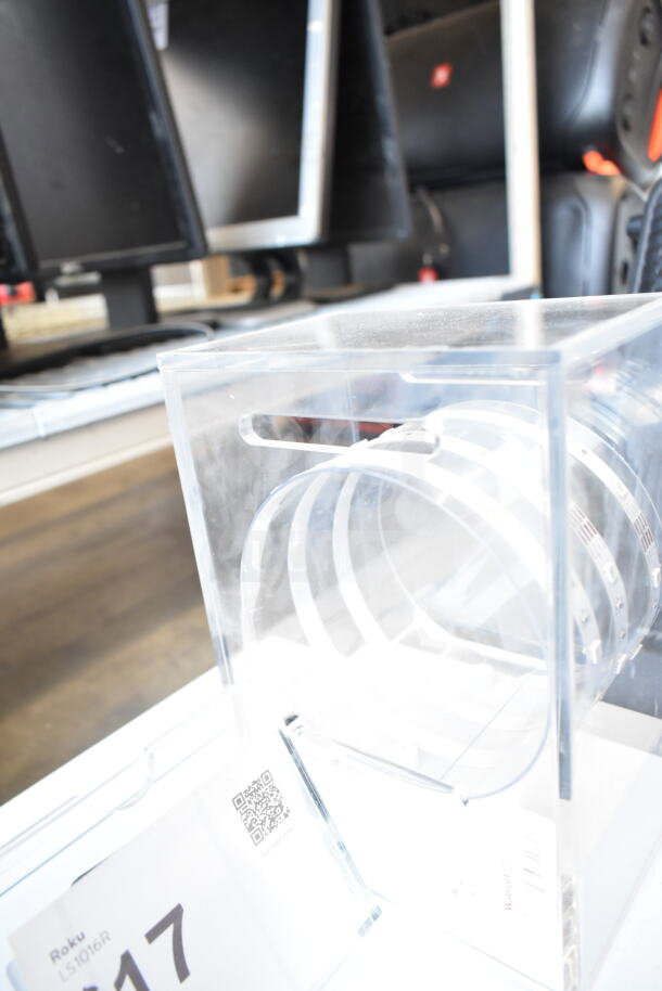 Clear acrylic display case labeled "Roku LS1016R," containing a coiled security band, showcasing the brand's camera securely.