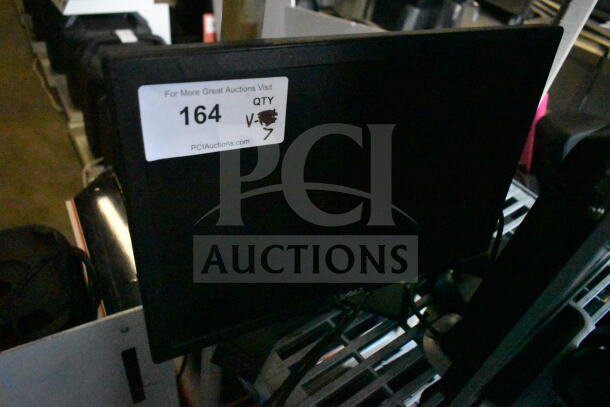 Seven various monitors, including a Dell, displayed in a warehouse. Label reads "164, Qty 7", indicating auction item quantity.