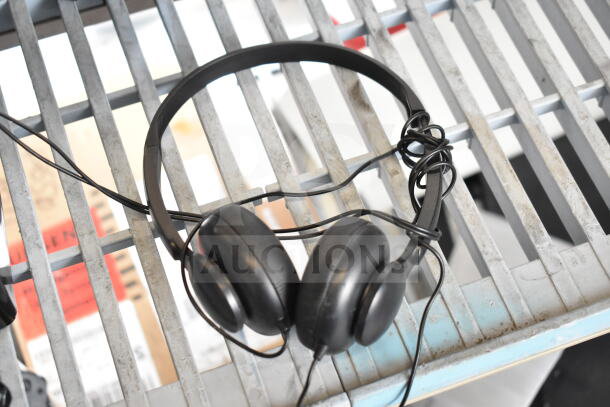 Three black over-ear headphones and a telephone, used, tangled cords, placed on a metal grate surface.