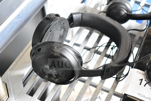 Black headphones on a metal shelf, featuring a logo on the earcup. Part of a lot including three headphones and a telephone.