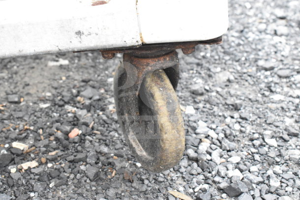 Rubbermaid pan transport rack caster, showing rust and wear on a gravel surface, indicating use and outdoor storage.