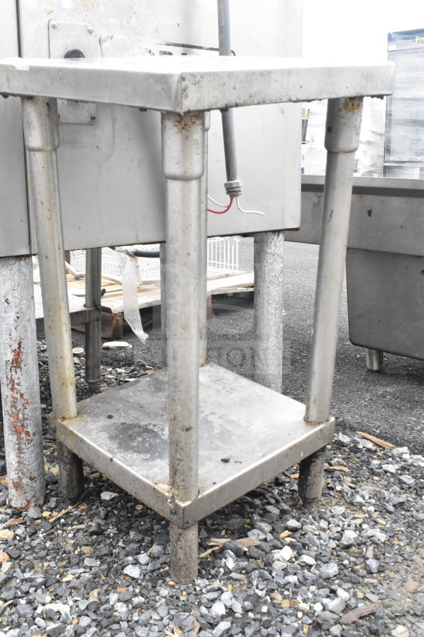 Stainless steel commercial table with under shelf, showing signs of wear and rust, stands outdoors on gravel.