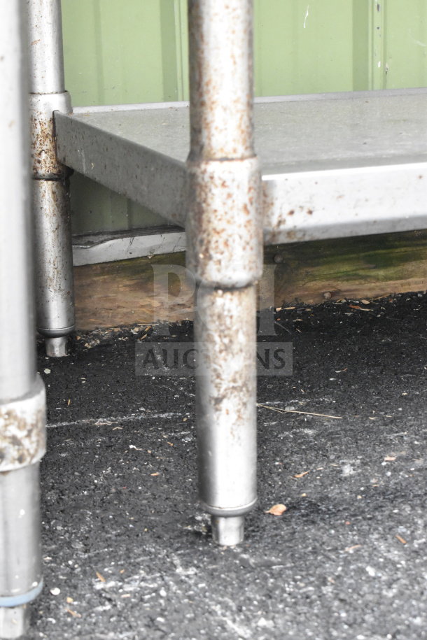 Commercial table with under shelf, metal legs show rust. Durable, ideal for heavy-duty use. Signs of wear visible.