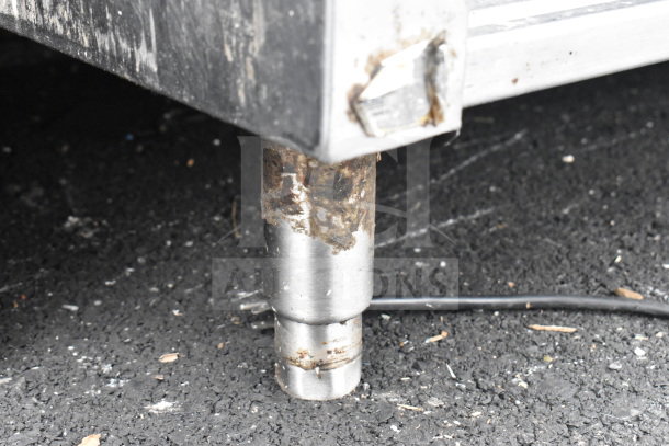 Stainless steel commercial table leg with rust and wear, visible under shelf, outdoors on asphalt surface.