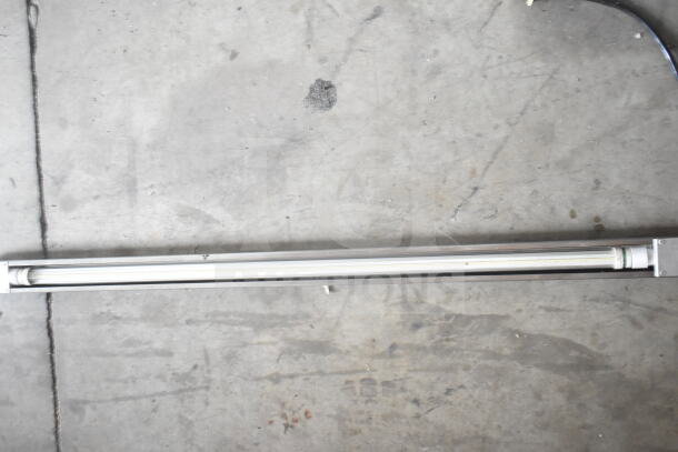 Industrial light fixture in metallic frame on concrete floor. Includes tube light, appears in good condition. No visible markings.