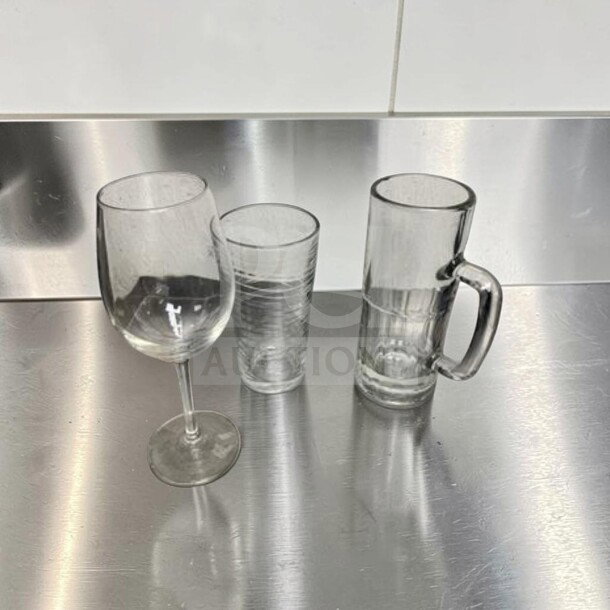 Three types of glassware on a stainless steel surface: a wine glass, a tumbler, and a beer mug. Suitable for restaurant use.